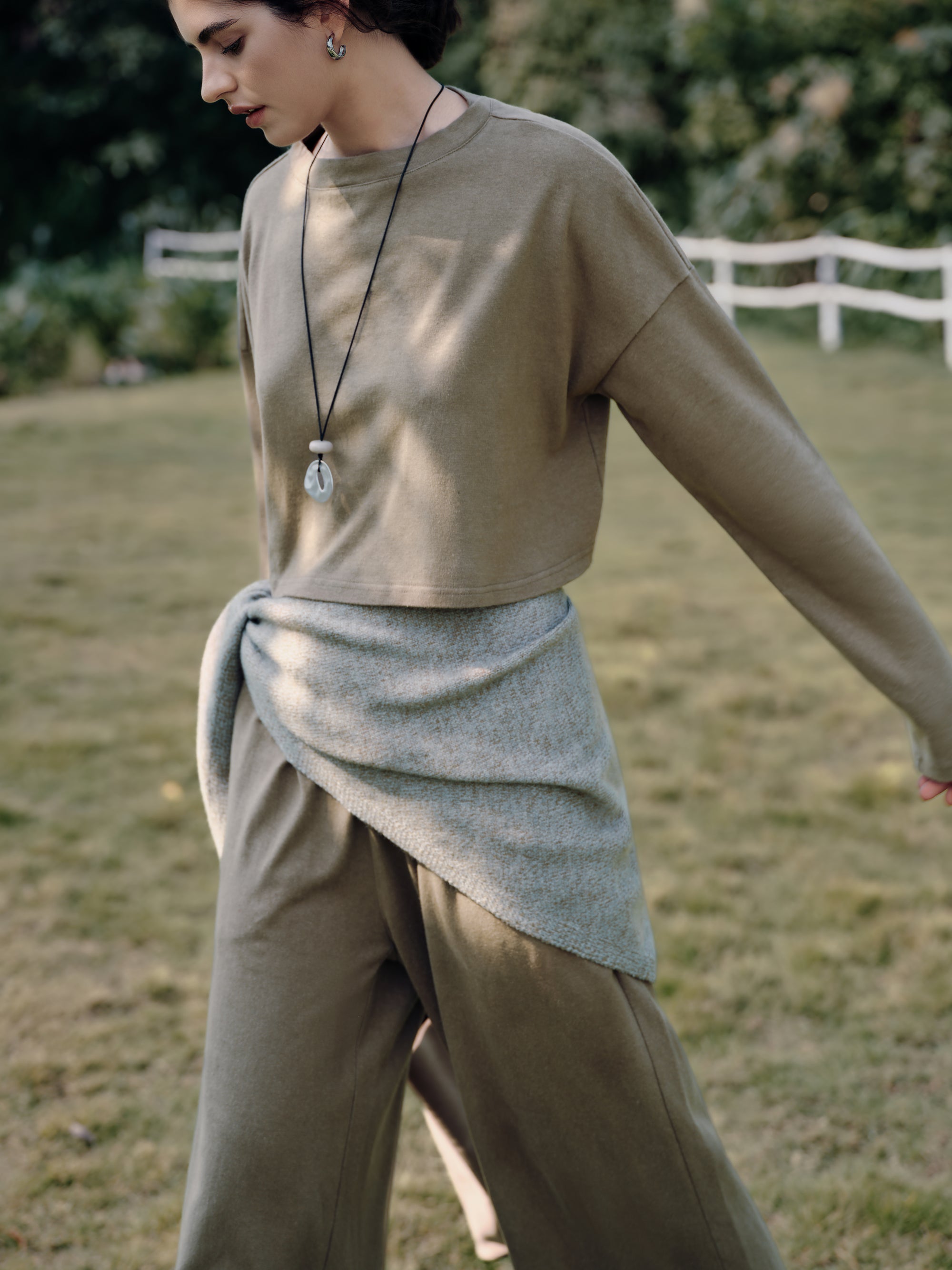 Model walking by a farmhouse gate wearing Juana Aera Organic Cotton Boxy Sweater olive with Aera Pull-on Pants olive (hemp–organic cotton blend), showing relaxed, breathable drape.