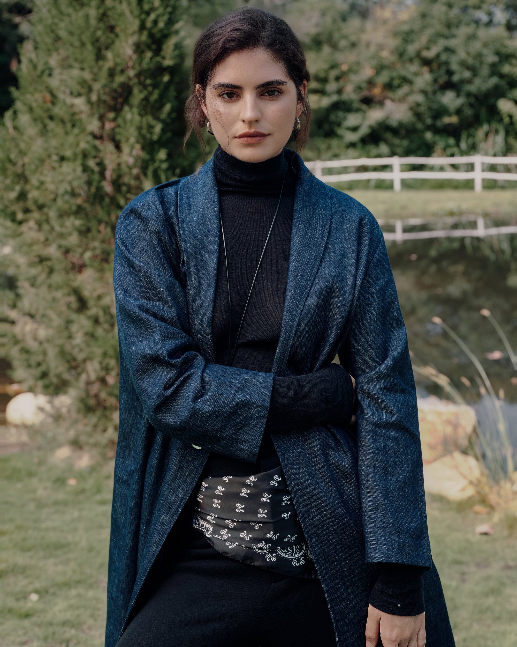 Garden standing shot of blue denim trench over black Ossa turtleneck and black high rise pants; clean longline silhouette and tailored drape.