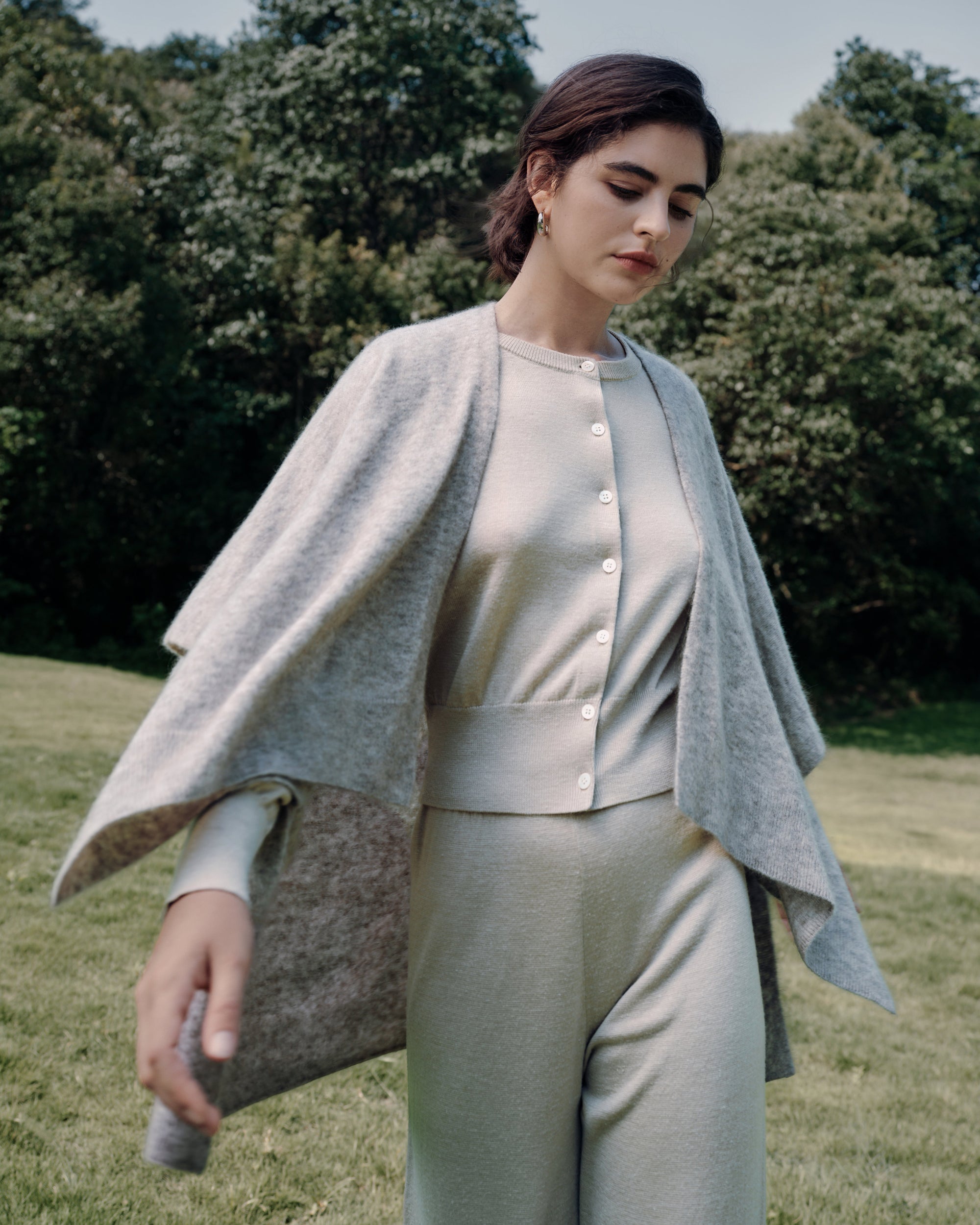 Model walking in a field wearing Liora Knitted Hemp Cardigan beige (merino–silk–hemp, shell buttons) with Nilo Knitted Hemp Pull-on Pants beige and Juana Cashmere Cape oatmeal (wool–cashmere–hemp), styled head to toe.