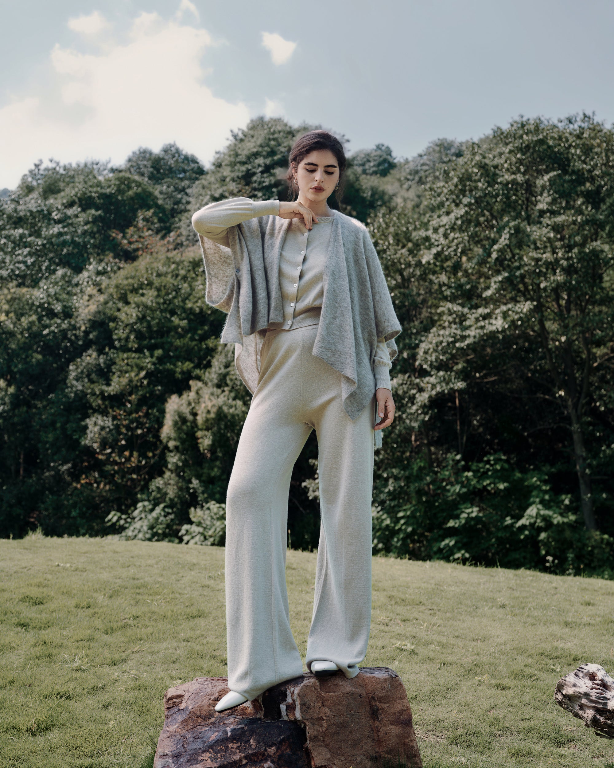 Garden standing shot of Liora beige layered under Cashmere Cape oatmeal with Nilo Pants beige; refined knit textures visible.