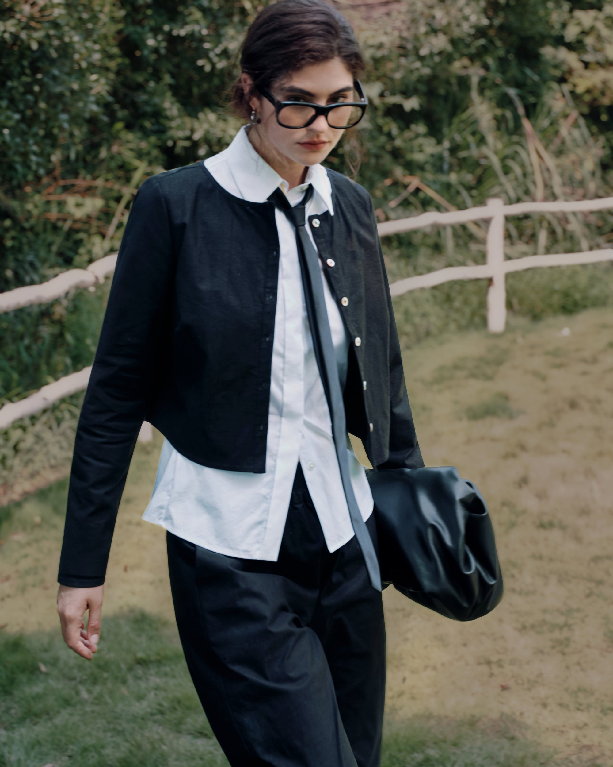 Model walking in a field wearing Juana Aella Long Sleeve Woven Top black (hemp–organic cotton stretch twill) over a white shirt, paired with Aella Mid Rise Relaxed Tapered Pants black; clean lines and breathable drape.