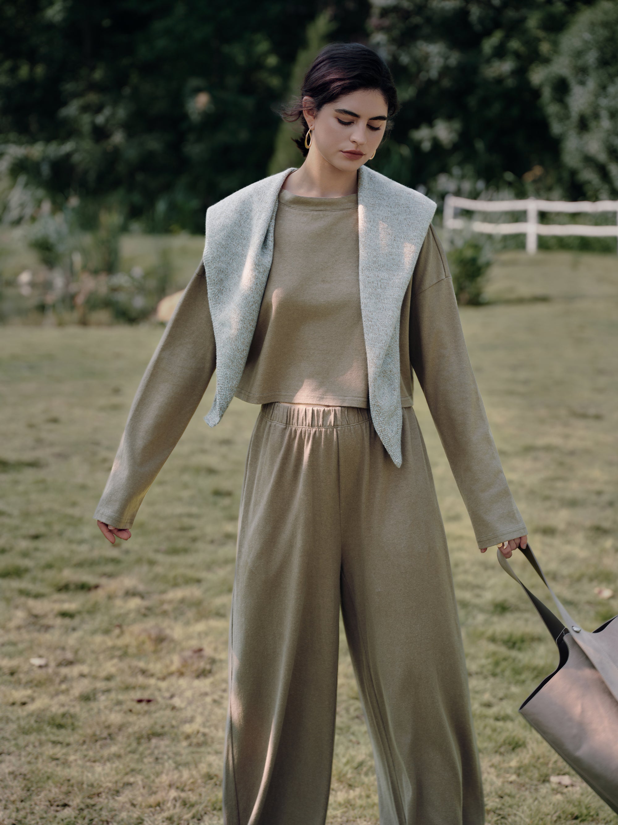 Portrait near a white fence of Aera Boxy Sweater olive and Pull-on Pants olive; soft volume and wide-leg flow in natural fibers.