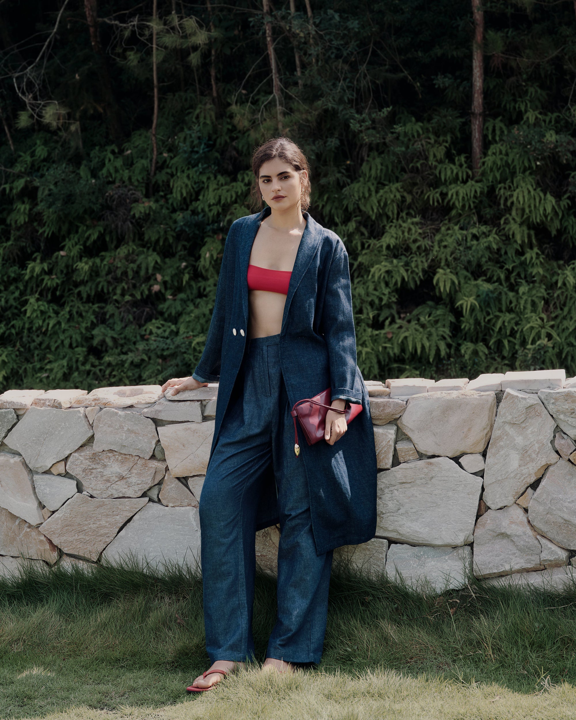 Leaning against a rock low wall in blue denim trench, indigo trousers, and red tube bra; minimalist, sustainable styling.