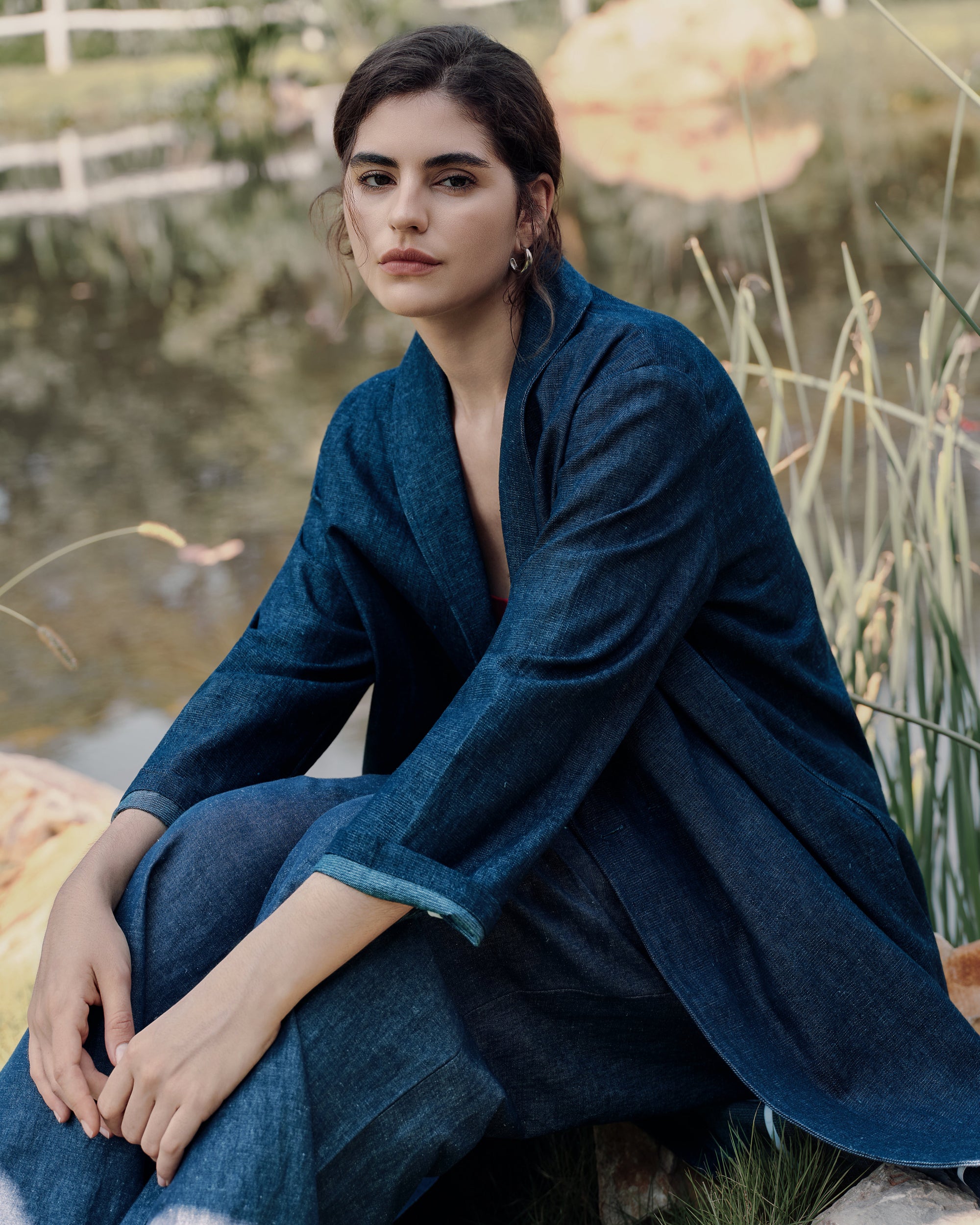 Sitting on a farmhouse fence wearing blue denim trench over black Ossa turtleneck with black classic high rise pants; elevated workwear mood.