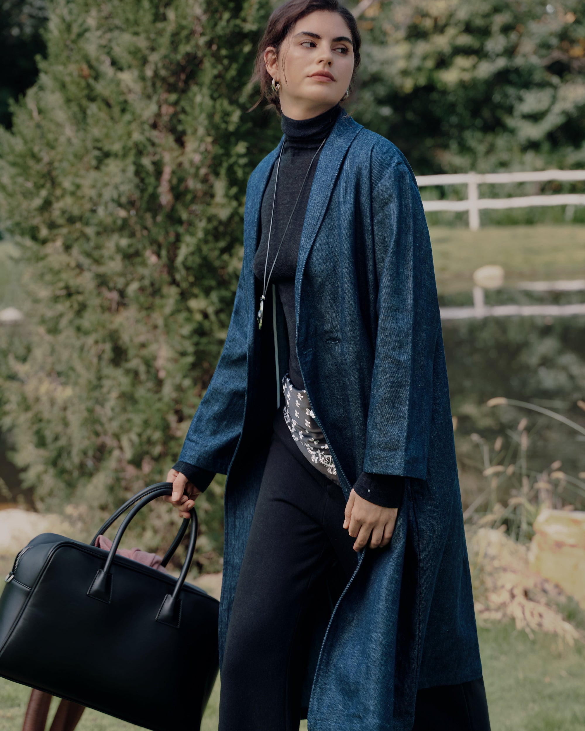 Three-quarter lakeside walk-away: front view of blue denim trench over black Ossa turtleneck and black classic high rise pants; elegant, seasonless layers.