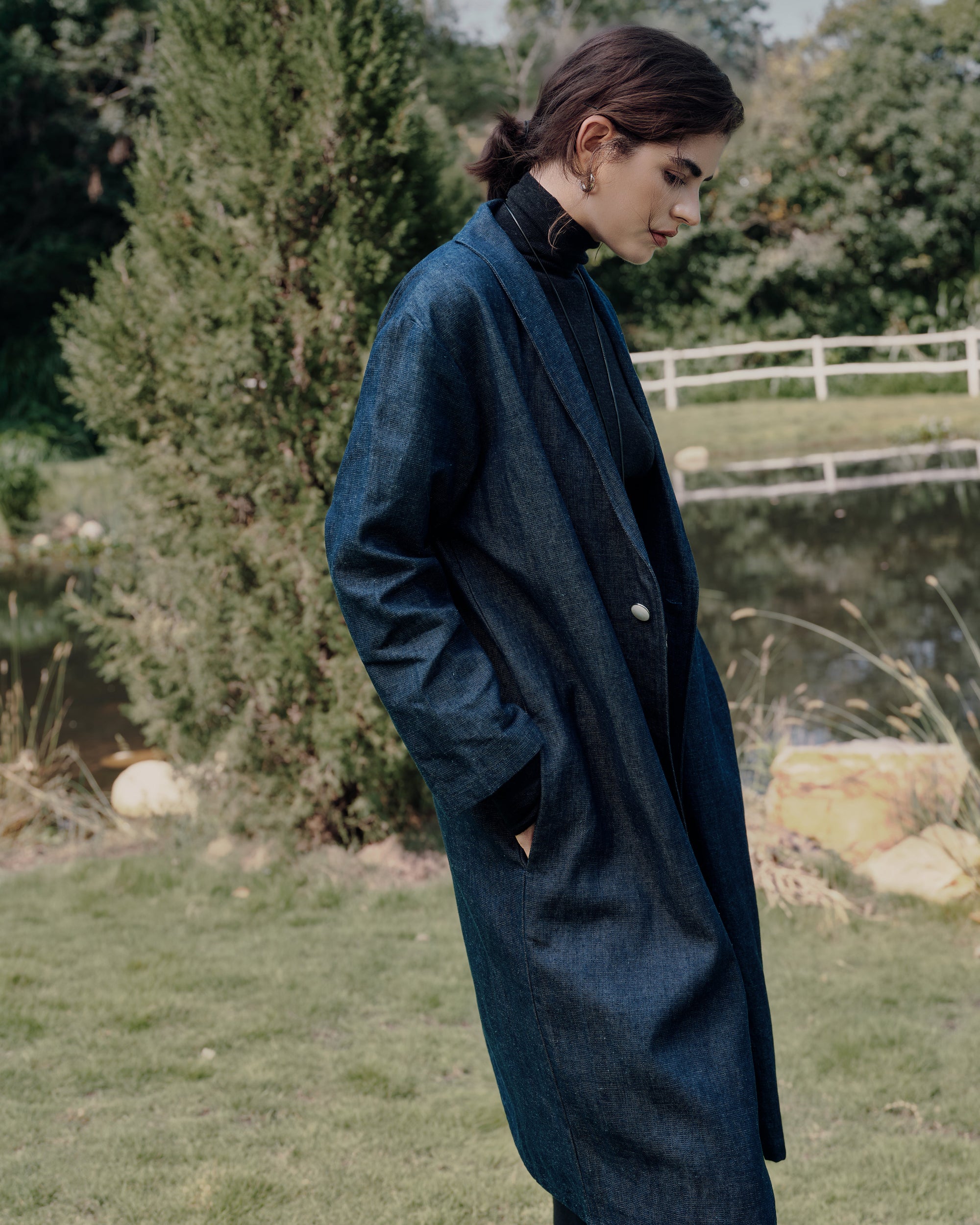 Side profile along a white-fence path: blue denim trench, showing pockets, black high rise pants; longline coat and clean trouser line.