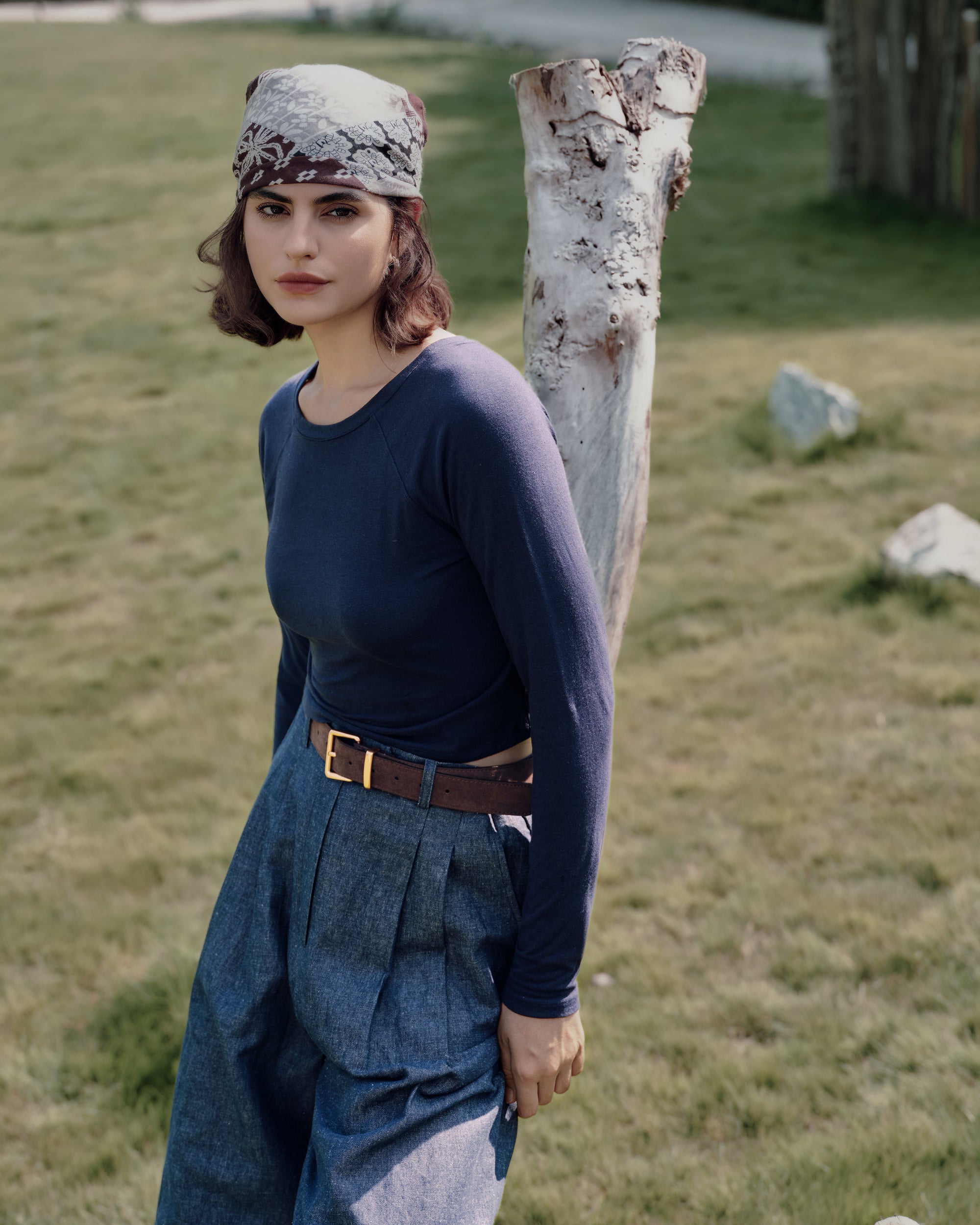 Leaning on a farmhouse log wearing indigo raglan tee and indigo high rise trousers; sustainable natural fibers, clean finish.