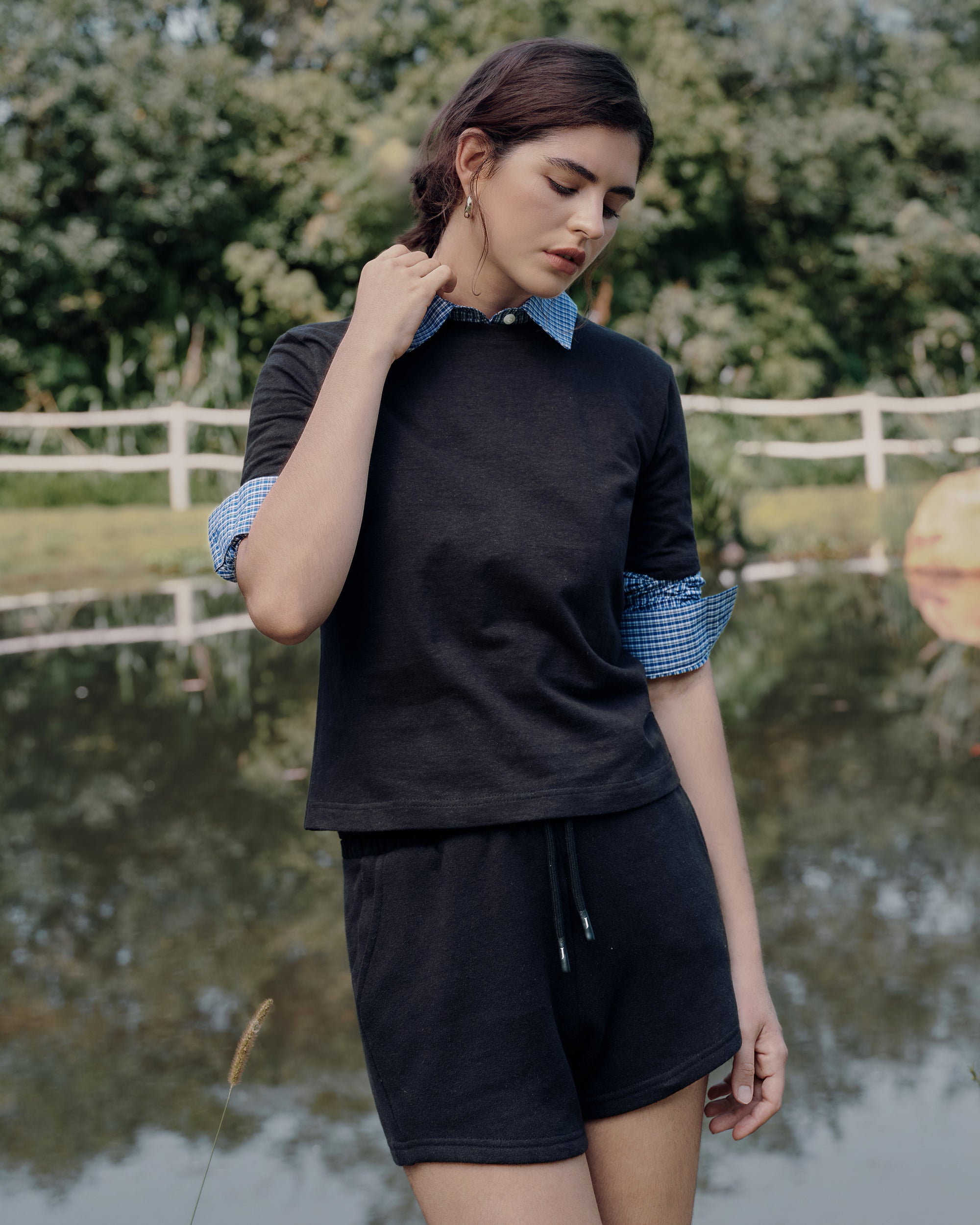 Model walking by a farmhouse gate wearing Juana Hemp Essentials Short Sleeve Tee white (hemp–organic cotton) over a blue shirt, paired with Hemp Essentials Fleece Shorts gray (hemp–organic cotton fleece).