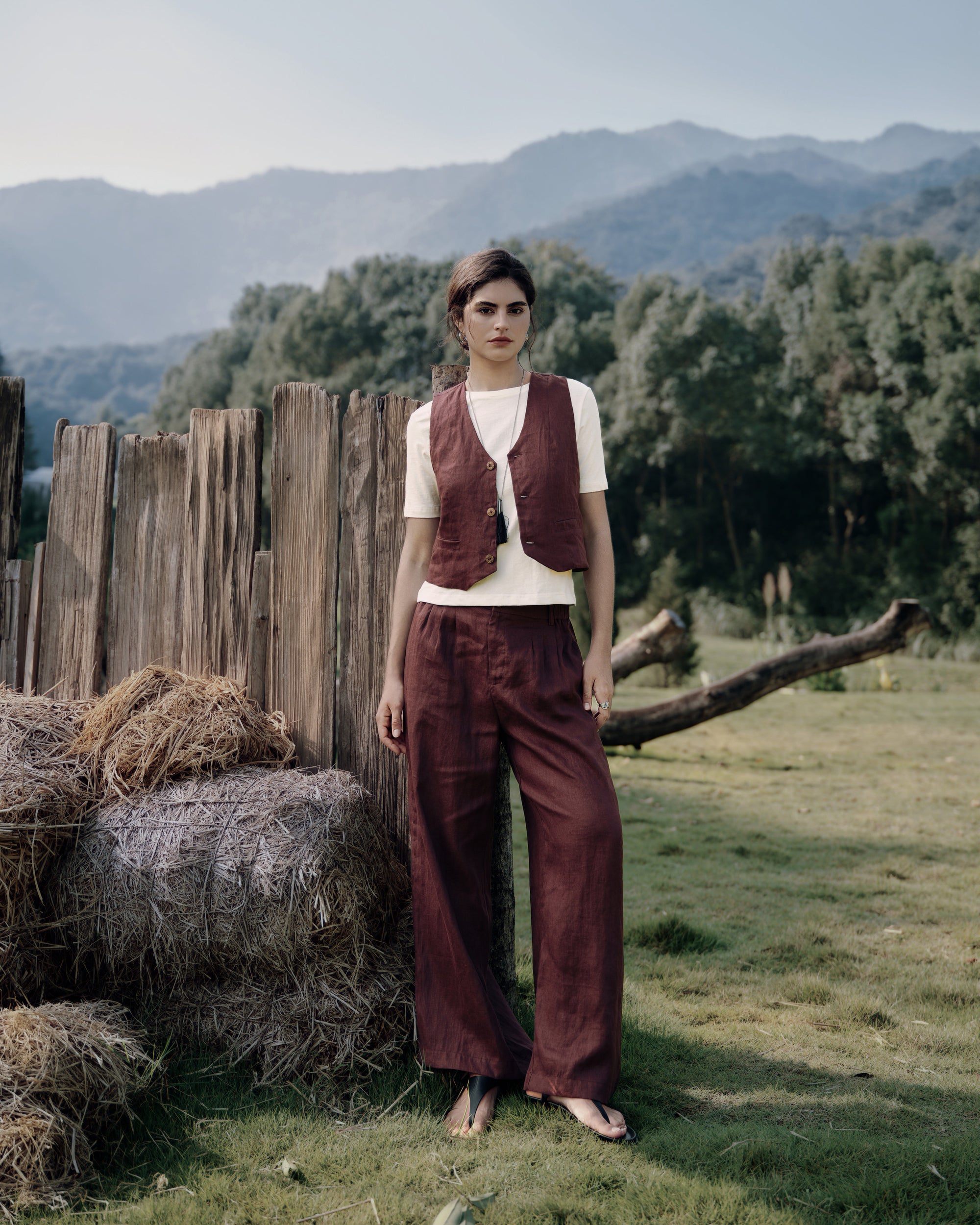 Standing at a barnwood fence in vest expresso with matching high rise pants expresso over white tee; tailored hemp set with clean lines.