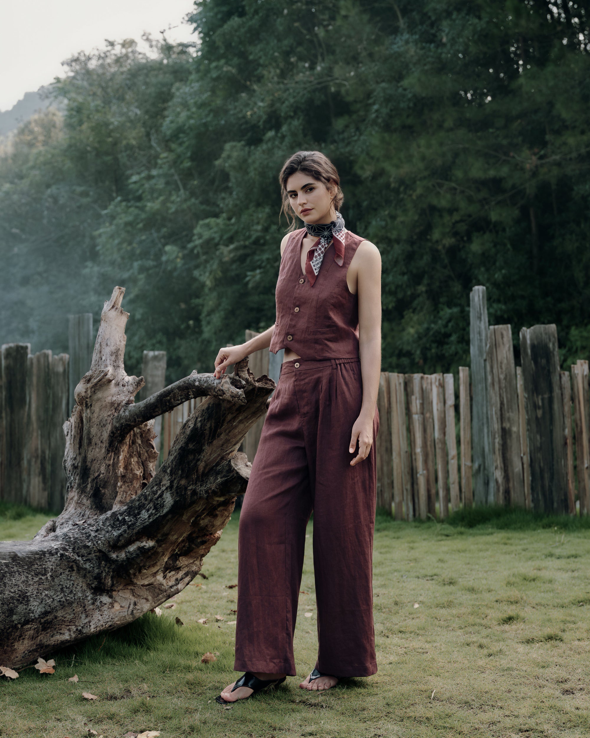 Model beside a large log on the farmhouse floor wearing Juana Hemp Tailored Vest expresso (100% hemp, corozo buttons) with Juana Hemp High Rise Pants expresso (100% hemp) over a white Hemp Essentials Short Sleeve Tee (hemp–organic cotton); grounded, natural styling.